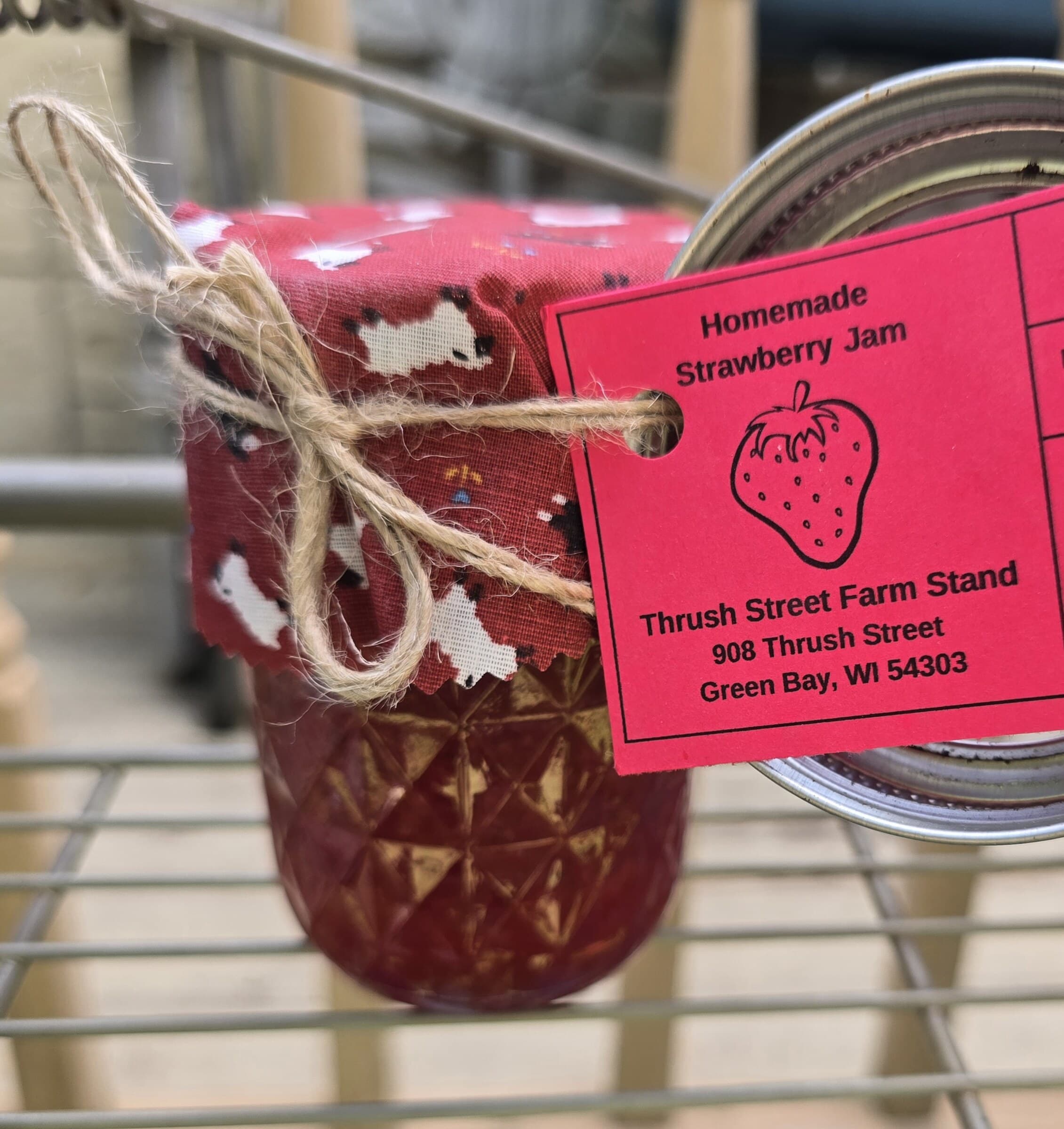 A jar of homemade strawberry jam with a pink fabric top and farm stand tag.